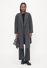 Cappotto in misto lana grigio con rever a punta, vestibilità rilassata, fessure laterali, abbinato a pantaloni in pelle nera e stivali con zeppa, che tiene una borsa con motivo.