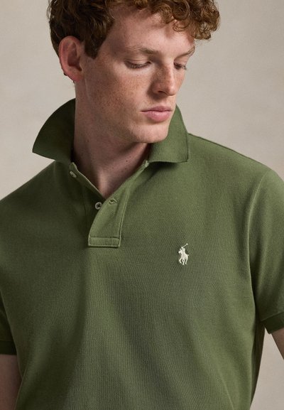 Oliivinvihreä pikeepaita, valmistettu teksturoidusta puuvillasta, jossa on kaksipainikkeinen nappilista ja vasemmassa rinnassa kirjailtu logo. Kauluksellinen malli.