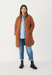 Giacca lunga imbottita marrone con colletto alto, zip frontale, tasche laterali, indossata sopra una camicia azzurro chiaro e jeans, abbinata a stivali neri.