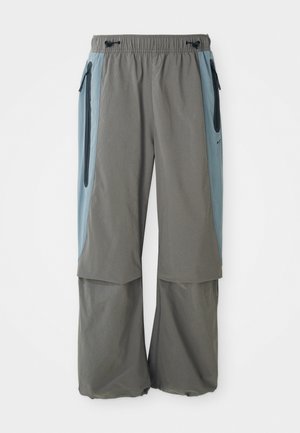 Grå utendørsbukser med bred benstil, med mørkeblå sidestykker og glidelåslommer. Laget av lett, pustende materiale.