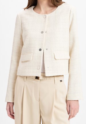 Veste en tweed beige clair avec un col rond, des boutons argentés et deux poches avant, associée à un pantalon beige.