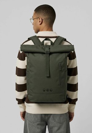 Junger Mann mit Brille, creme-braun gestreiftem Pullover, grauer Jeans und dunkelgrünem Rolltop-Rucksack, von hinten auf einfarbigem Hintergrund.