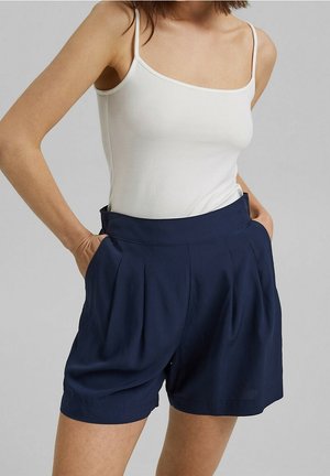 Femme portant un top blanc à fines bretelles et un short bleu marine taille haute plissé, les mains dans les poches, devant un fond uni.