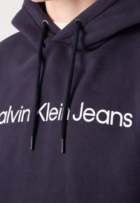 Mörkblå lurvig huvtröja som är gjord av mjuk bomullsblandning, med en framträdande vit "Calvin Klein Jeans" logotyp och vävda svarta dragsnören med metallaglets.