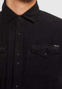 Camisa de pana negra con cuello abotonado, que presenta dos bolsillos en el pecho con botones de presión y un pequeño logo blanco "REPLAY" como detalle.