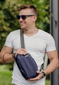 Navy modrý křížový kabelka s hladkou texturou a zipem, kterou drží osoba oblečená ve fitted bílém tričku a slunečních brýlích na venkovním místě.