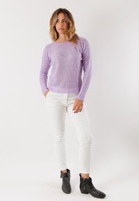 Donna in piedi, che indossa un maglione lavorato a maglia lilla, pantaloni bianchi e stivaletti neri, su uno sfondo chiaro e semplice.