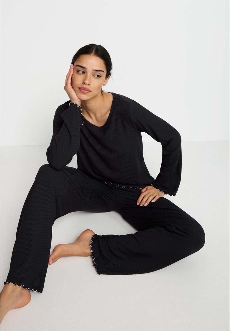 Ensemble de détente noir à côtes comprenant un haut à manches longues avec un décolleté en scoop et un pantalon avec des ourlets en scallop. Tissu confortable et coupe décontractée.
