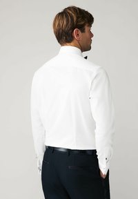 Weißes Hemd mit klassischem Kragen, Knopfmanschetten mit schwarzen Akzenten, getragen mit dunklen Hosen. Glatte, eng anliegende Stofftextur.