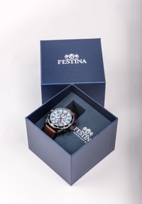 Rudas odinis laikrodis mėlyname dėžutėje. Jame yra šviesiai melsvas ciferblatas, sidabrinės ir juodos detalės bei keli ciferblatai su raudonomis detalėmis.