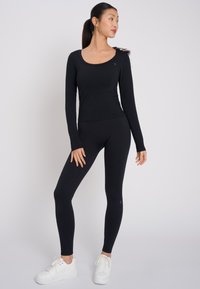 Schwarzes Langarmoberteil und Leggings aus strukturiertem, dehnbarem Stoff. Mit einem Rundhalsausschnitt und subtilen Logodetails, kombiniert mit weißen Sneakers.