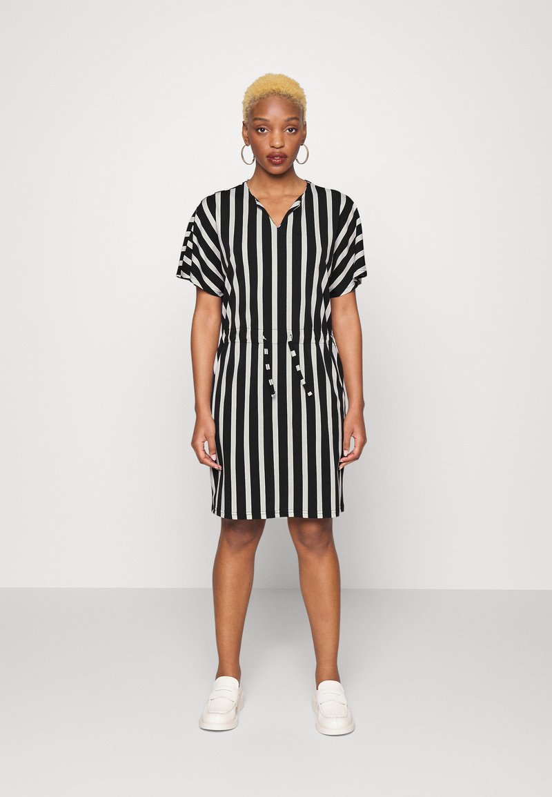 Vestido às riscas preto e branco com decote em V, mangas curtas, cintura com cordão e comprimento até ao joelho, combinado com mocassins de cores claras.