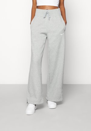 Personne portant un pantalon de survêtement gris clair à jambes larges avec le logo Nike, des baskets blanches et un crop top blanc, debout les mains dans les poches.