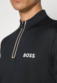 Svart zip-up tröja i strukturerad tyg, med en guldutrustad dragkedja och vit "BOSS" logotyp på bröstet.