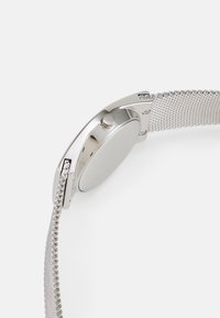 Zegarek z siatki ze stali nierdzewnej z okrągłą srebrną kopertą, minimalistyczny design i gładkie wykończenie. Posiada boczną koronkę do regulacji.