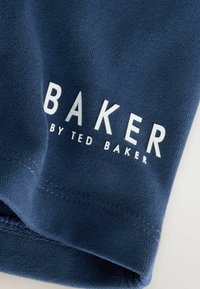 Zbliżenie na granatowy materiał z białym nadrukiem tekstu "BAKER BY TED BAKER" i widocznym przeszyciem wzdłuż złożonej krawędzi.