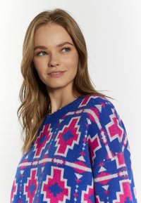 Pulover cu model strălucitor albastru, prezentând forme geometrice în roz și alb. Guler rotund și textură moale, evidențiind un design intricată de tricot.