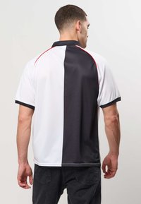 Homme vu de dos portant un polo à manches courtes avec un design vertical noir et blanc divisé et des finitions rouges aux épaules.