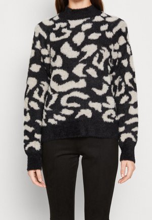 Woman wearing black and white fuzzy animal print sweater with long sleeves and high neck, paired with black pants.