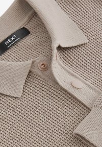 Camisa polo de punto beige con un patrón texturizado, que presenta un cuello y dos botones hechos de un material liso a juego. Etiqueta de la marca visible.