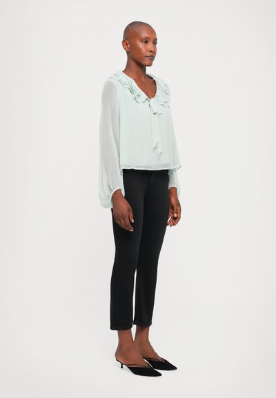 Light green blouse with ruffled neckline and bell sleeves, paired with black fitted pants and pointed black heels.