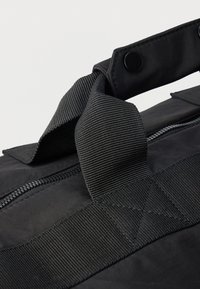 Μαύρη τσάντα με υφασμάτινο λουράκι με υφή και λείο φερμουάρ, κατασκευασμένη από ανθεκτικό ύφασμα με ματ φινίρισμα, που ξεχωρίζει από τη κεντρική ραφή και το κούμπωμα με κουμπί.