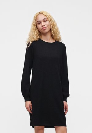 Jonge vrouw met lang krullend blond haar, die een eenvoudige zwarte jurk met lange mouwen draagt, staand voor een witte achtergrond.