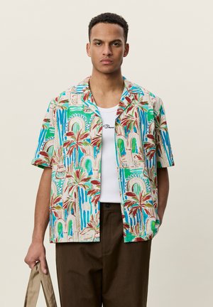 Homme portant une chemise tropicale colorée à manches courtes par-dessus un débardeur blanc et un pantalon marron, tenant un sac beige dans sa main gauche.