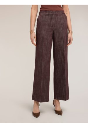 Mujer vistiendo pantalones anchos de pierna con textura marrón, top marrón sin mangas, brazaletes dorados y tacones marrones de punta puntiaguda sobre un fondo liso.