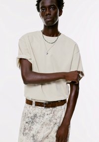 Beige, oversized póló, felgöngyölt ujjal, mintás világos nadrággal párosítva. A modellen zöld gyöngy nyaklánc és barna öv található.