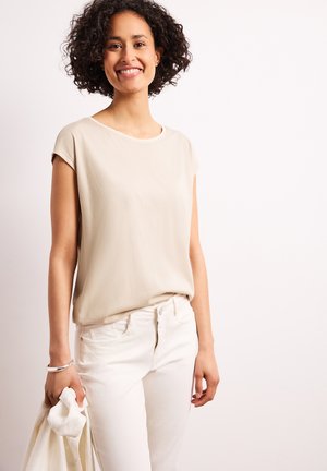 Smilende kvinde med krøllet hår iført en beige ærmeløs top og hvide bukser, der holder en lys jakke i venstre hånd.