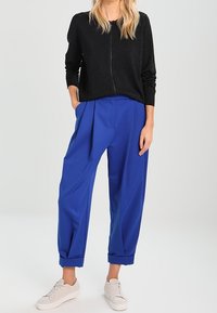 Maglione nero con texture e cerniera, abbinato a pantaloni blu plissettati e sneakers bianche. I pantaloni hanno risvolti e una vestibilità comoda.