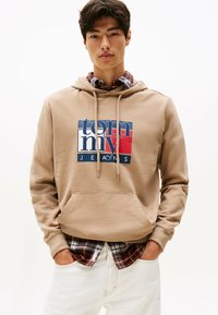 Giovane uomo che indossa un cappuccio beige con il logo Tommy Jeans sopra una camicia a scacchi rossa e nera e pantaloni bianchi, con le mani in tasca, su uno sfondo neutro.