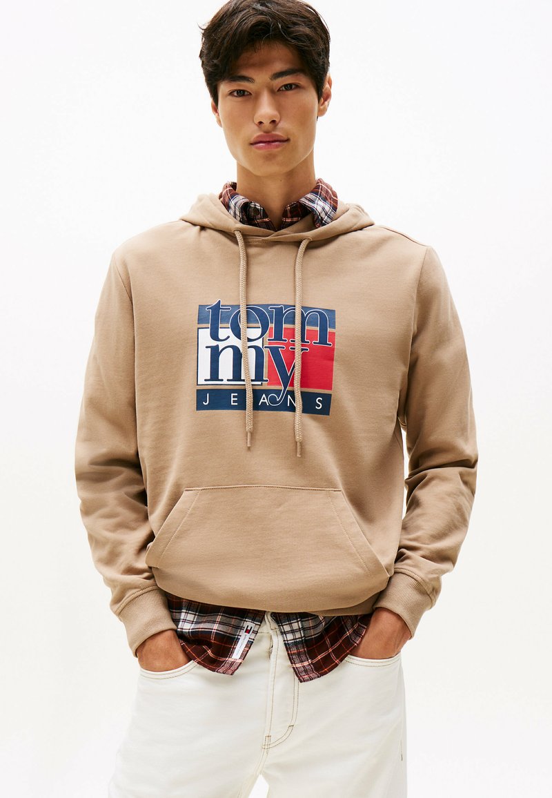 Giovane uomo che indossa un cappuccio beige con il logo Tommy Jeans sopra una camicia a scacchi rossa e nera e pantaloni bianchi, con le mani in tasca, su uno sfondo neutro.