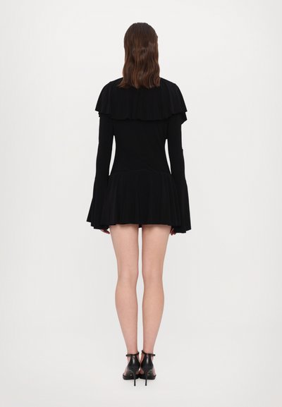 Zwarte jurk van gladde stof met gelaagde ruches bij de schouders en een uitlopende rok. Gestyled met zwarte hakken.