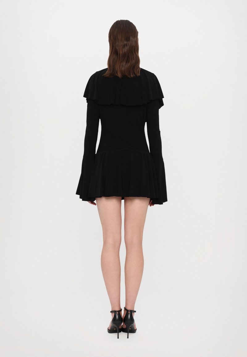 Zwarte jurk van gladde stof met gelaagde ruches bij de schouders en een uitlopende rok. Gestyled met zwarte hakken.