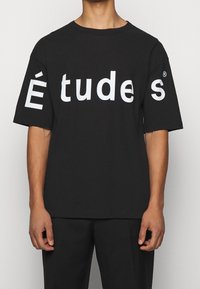 Musta puuvillainen t-paita, jossa on ylisuurikokoinen istuvuus ja suuri valkoinen teksti "É tud es" paidan edessä. Lyhyet hihat ja pyöreä pääntie.