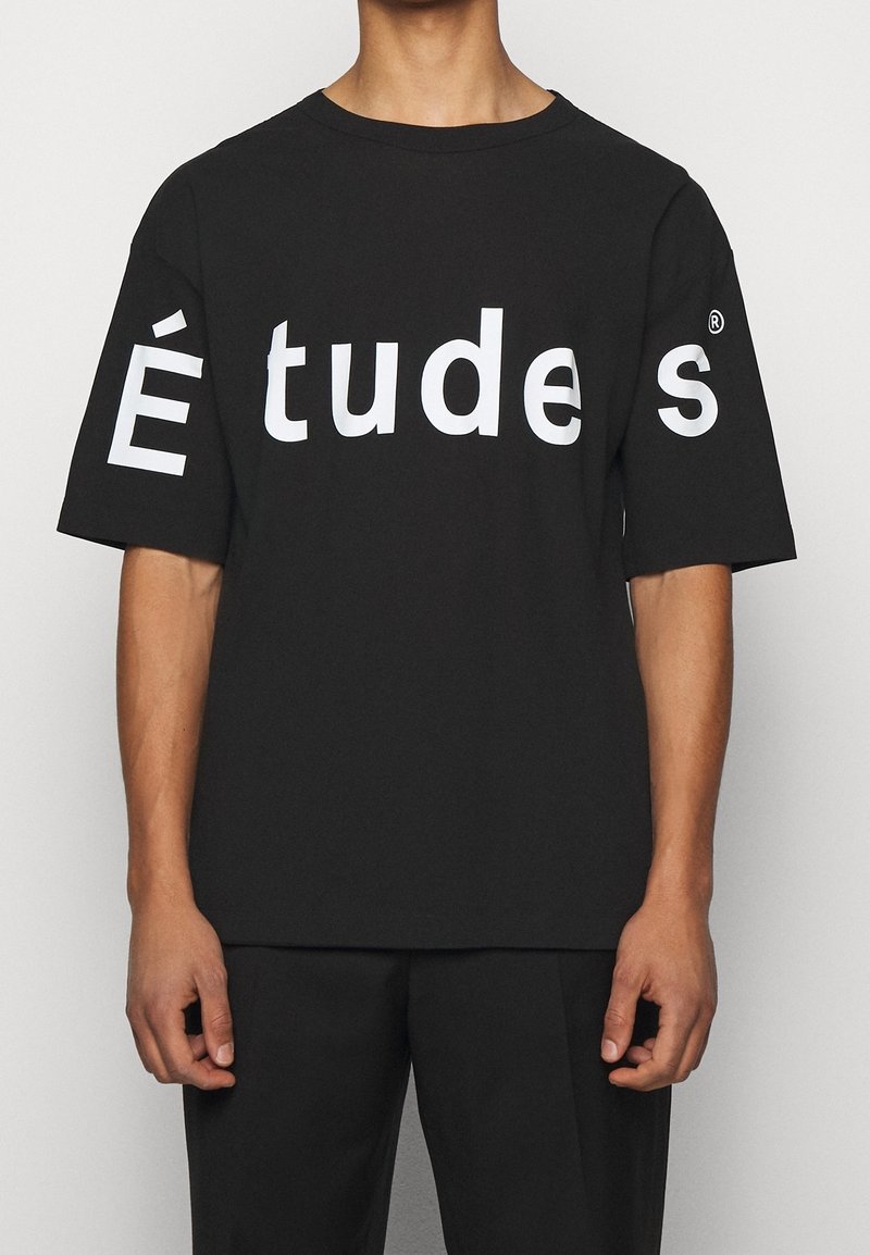 Musta puuvillainen t-paita, jossa on ylisuurikokoinen istuvuus ja suuri valkoinen teksti "É tud es" paidan edessä. Lyhyet hihat ja pyöreä pääntie.