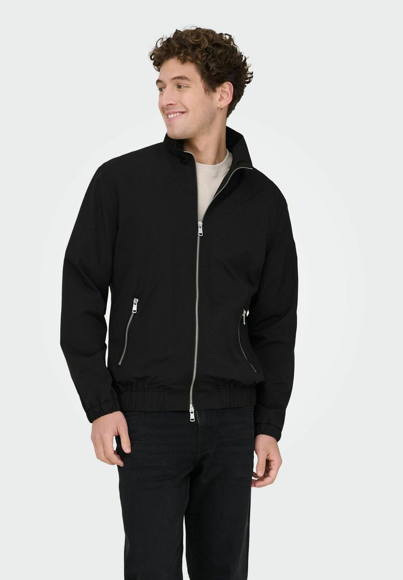 Schwarze leichte Jacke mit Frontreißverschluss, zwei Seitentaschen, elastischem Bund und Bündchen, ausgestattet mit hohem Kragen und glatter Textur.