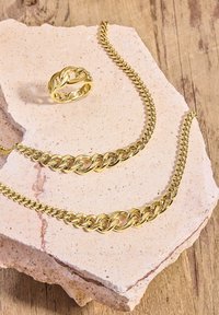 Collana, bracciale e anello in oro placcato su una superficie di pietra chiara. Presentano maglie intrecciate con una finitura lucida, creando un aspetto splendente.