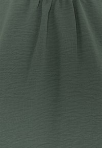Tessuto di un verde scuro, testurizzato con una leggera lucentezza e lievi pieghe, caratterizzato da morbide cadute e un dettaglio arricciato nella parte superiore.