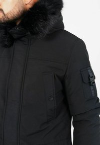 Svart parka-jacka med en huva i konstpäls, dragkedja och tryckknappar, flera fickor och en mjuk yta. Accenter på ärmen.