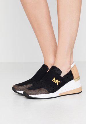 Stopy w czarnych slip-on sneakersach z złotym logo "MK", białymi podeszwami i beżowymi akcentami na jednolitym jasnym tle.