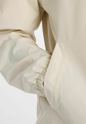 Hellbeigefarbener Jackenärmel mit elastischer Manschette und teilweise in einer Seitentasche versteckte Hand auf einfarbigem Hintergrund.