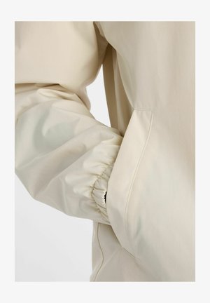 Manica di giacca beige chiaro con polsino elastico e mano parzialmente infilata in una tasca laterale su sfondo uniforme.