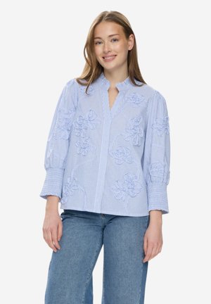 Jonge vrouw met lichtbruin haar, gekleed in een lichtblauw gestreept blouse met bloemrijke borduurwerk en een blauwe spijkerbroek, glimlachend en staand.