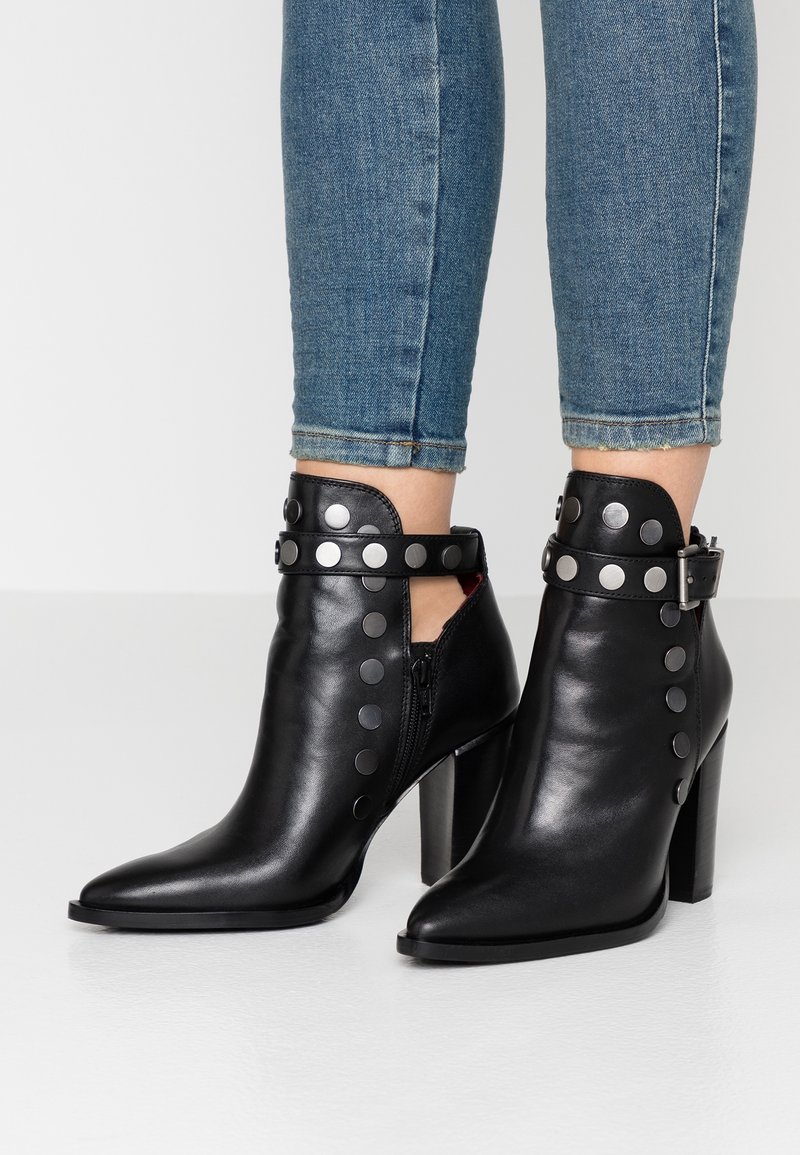 Bottes basses en cuir noir avec bouts pointus, talons épais, clous argentés et sangles à boucle portées avec un jean bleu.