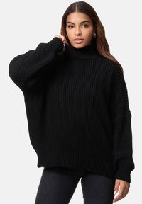 Schwarzer oversized Strickpullover mit gerippter Textur, hohem Kragen und langen Ärmeln. Kombiniert mit dunklen Jeans, die eine lockere Passform haben.