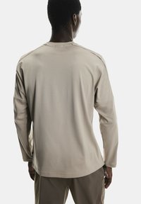 Cămașă cu mâneci lungi de culoare bej deschis, având un material texturat. Cusăturile sunt vizibile pe umeri și pe părțile laterale, cu un tiv rotunjit.