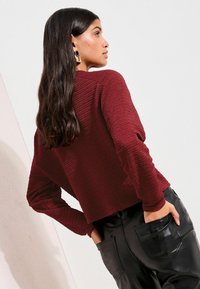 Pulover texturat bordo cu mâneci lungi, asortat cu pantaloni negri cu talie înaltă și buzunare mari. Finisaj neted al pantalonilor.